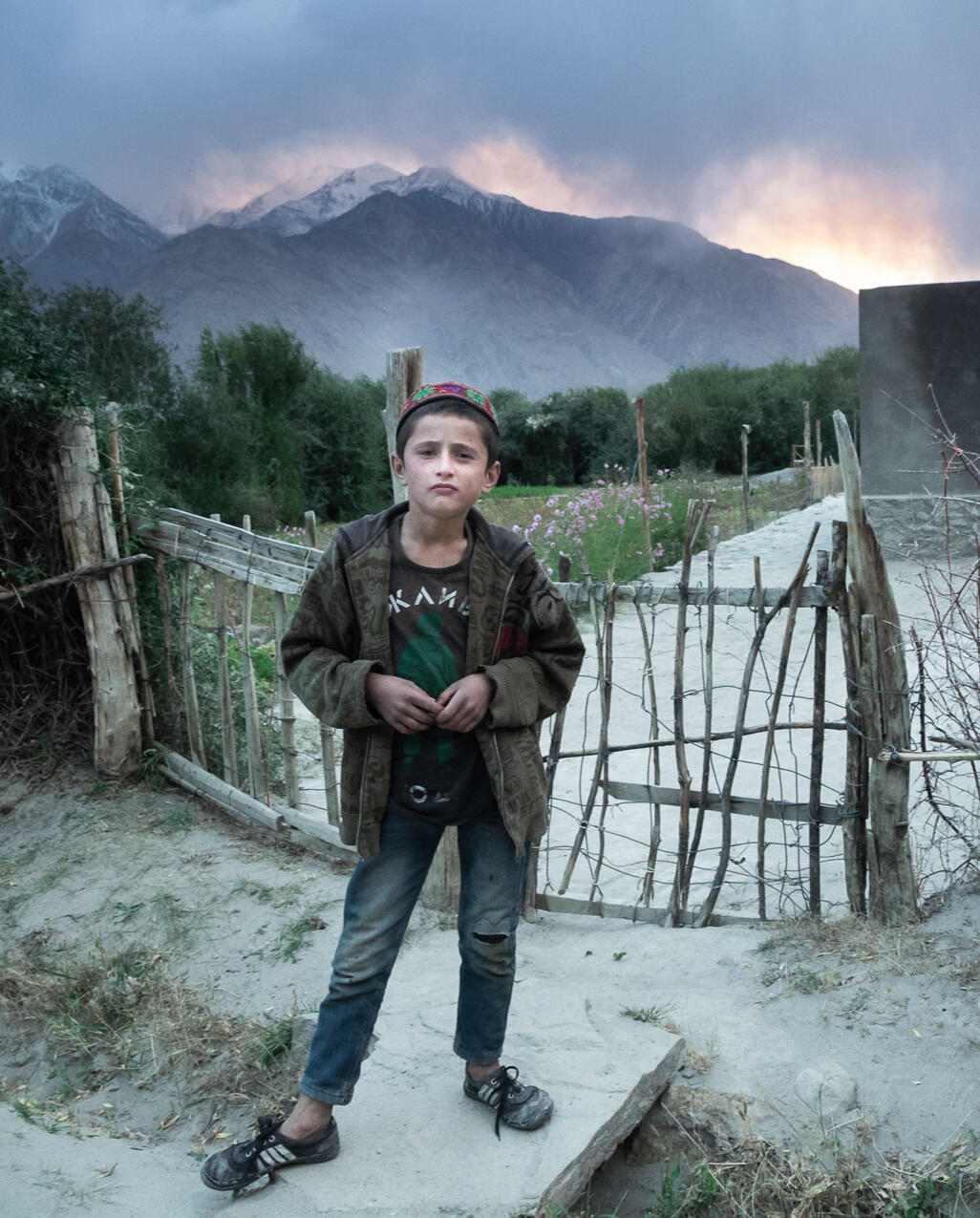 frontier-discovery-project-boy-village-home.jpg Village boy stands in front of fence at home with trees and mountains behind him